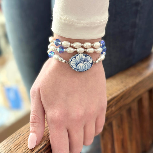 May include: A three-strand bracelet set. The top two strands feature white pearls and blue crystal accents. The bottom strand has a blue and white floral cameo. The bracelets are worn on a wrist.