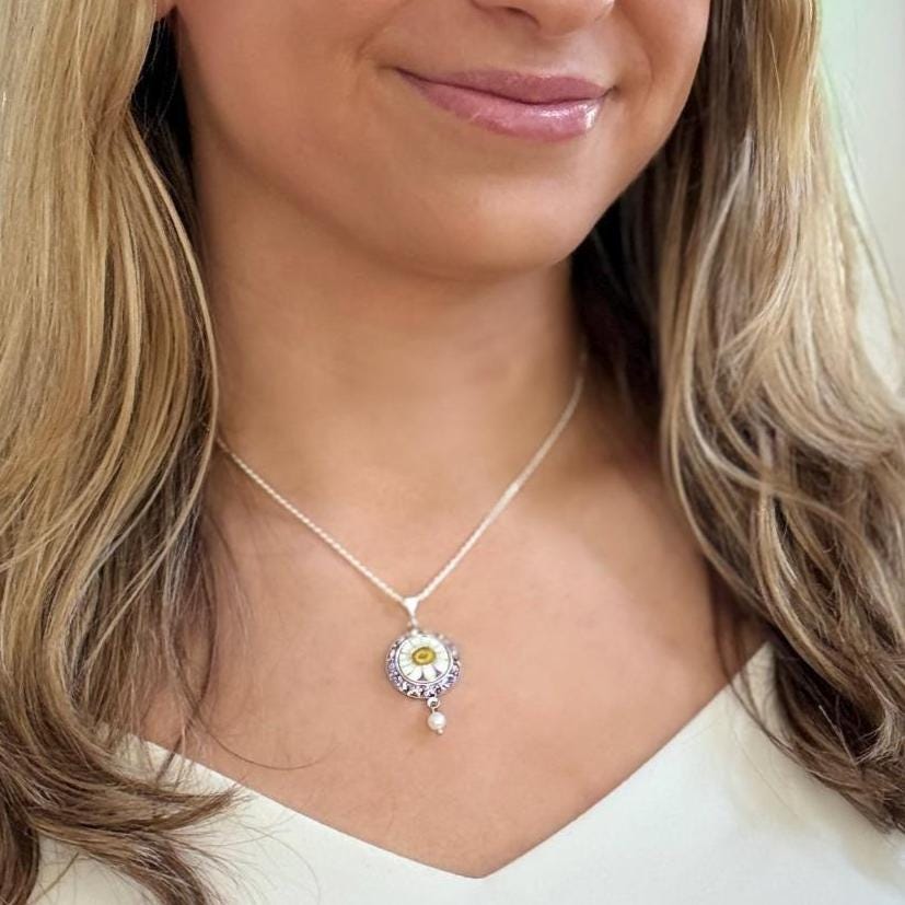 May include: A silver necklace with a daisy pendant. The pendant features a yellow center, white petals, and a small pearl drop. The necklace is worn by a person with blonde hair and blue eyes.