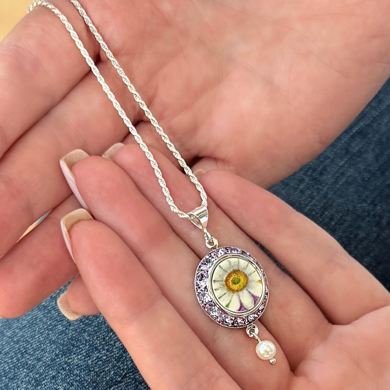 May include: Silver necklace with a daisy pendant. The pendant features a real daisy encased in a clear resin, surrounded by purple crystals. A small pearl dangles below the pendant. The necklace is held in a person's hands.