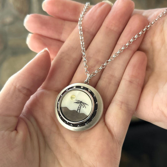 May include: Silver-tone pendant necklace featuring a round locket with a beach scene. The locket has a black border with small, dark accents. Inside, a palm tree and sun are depicted against a white and gray background. The chain is a delicate link style.