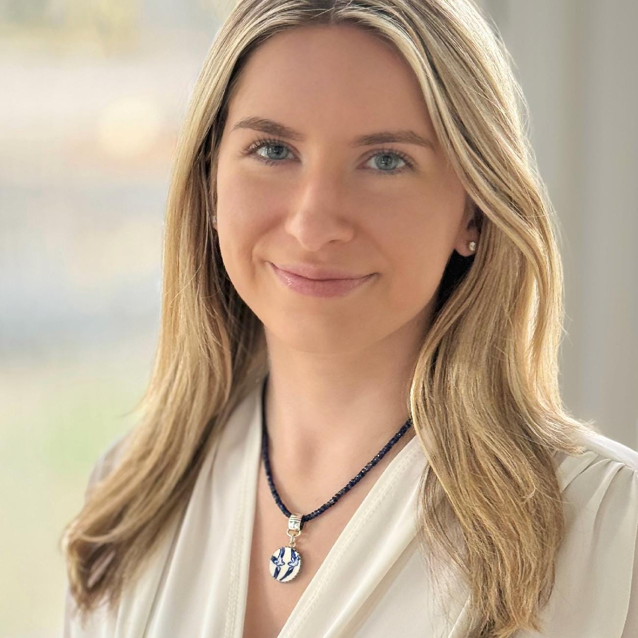 May include: A dark blue beaded necklace with a round pendant. The pendant has a white and blue design. The necklace is worn by a person with blonde hair and a white blouse.