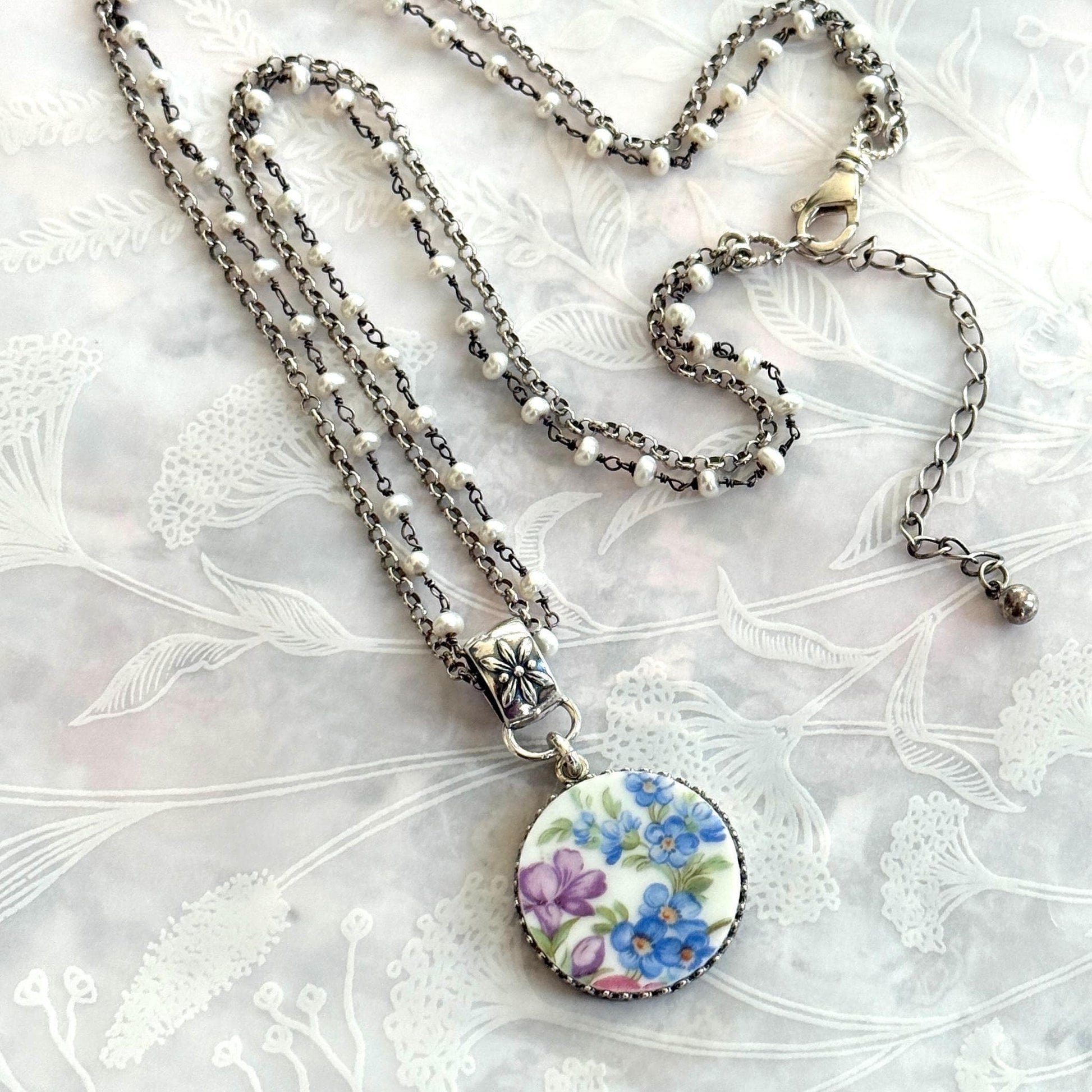 May include: A silver-toned necklace with a floral pendant. The pendant features a round, white ceramic design with blue and purple flowers. The necklace has multiple strands of small white beads and a decorative clasp.