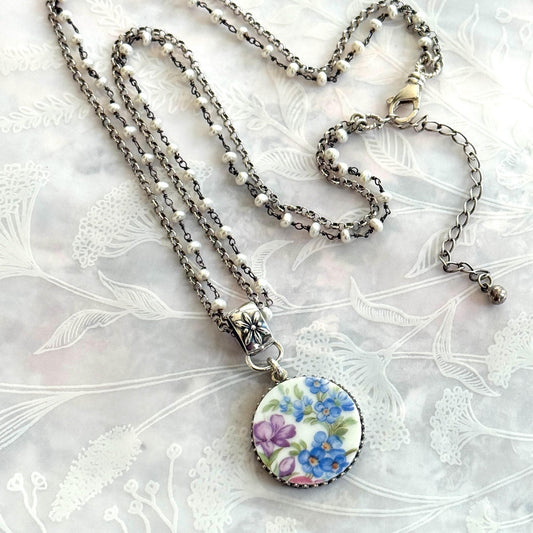 May include: A silver-toned necklace with a floral pendant. The pendant features a round, white ceramic design with blue and purple flowers. The necklace has multiple strands of small white beads and a decorative clasp.