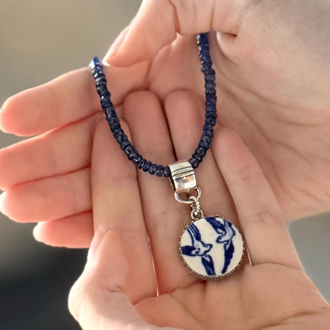 May include: A handcrafted necklace featuring a dark blue beaded chain and a circular pendant. The pendant has a white background with a blue bird design and a silver-colored metal frame. The necklace is held in open hands.