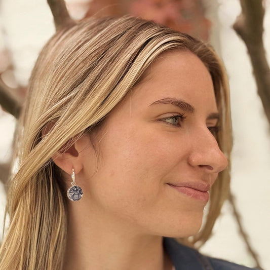 May include: Silver hoop earrings with a round, blue and white pendant. The pendant features a geometric design with a central square and surrounding details. The earrings are worn by a person with blonde hair.