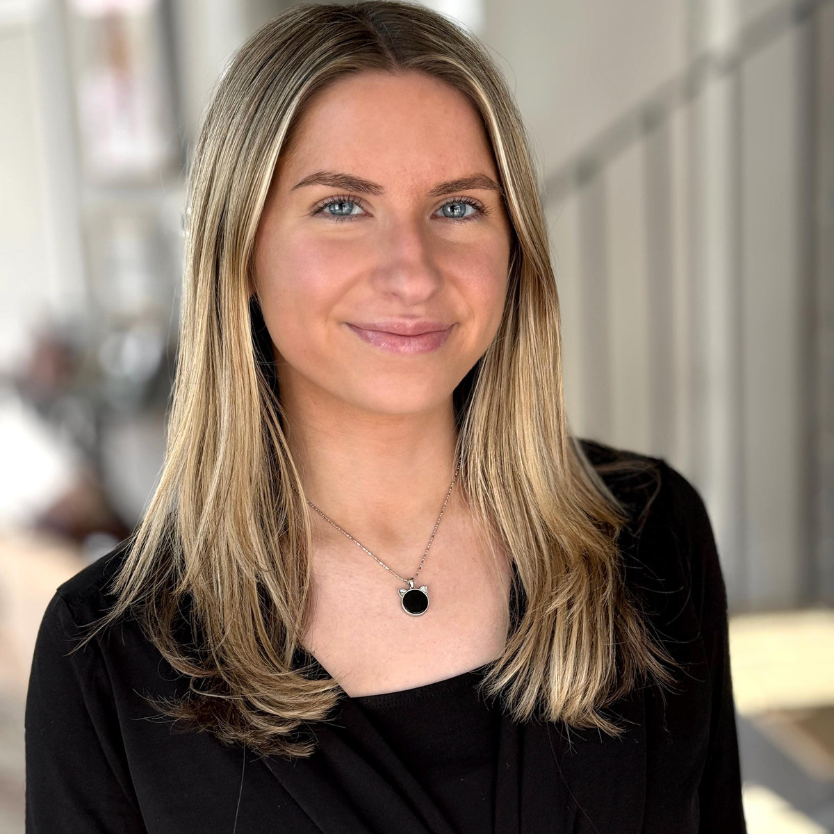 May include: A woman with long blonde hair and blue eyes is wearing a black top and a silver necklace with a black pendant. The background is blurred, with a focus on the woman's face and upper body.