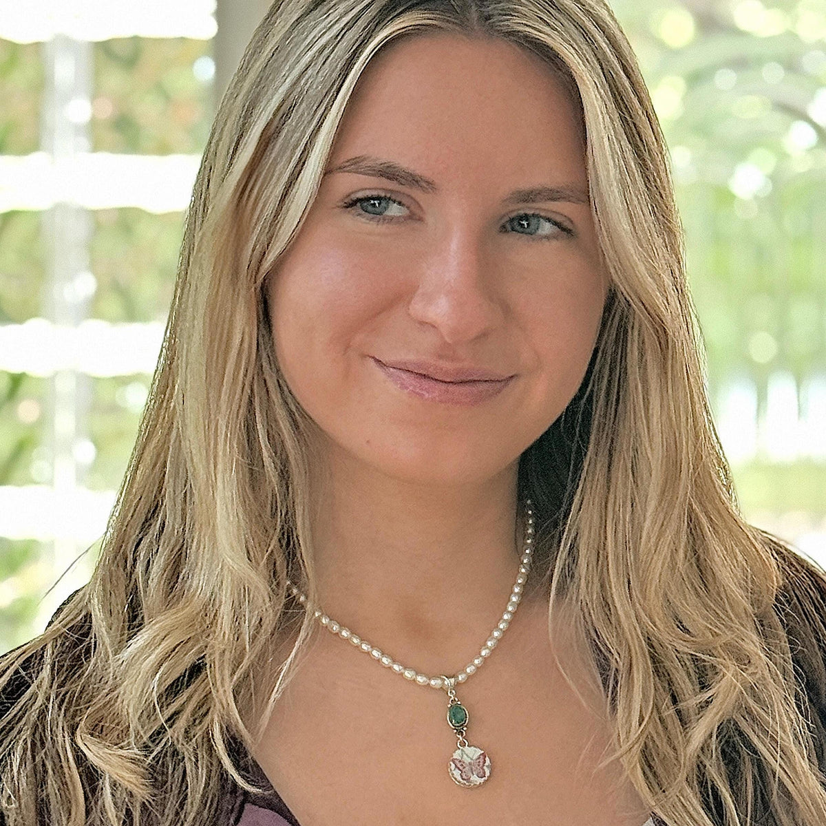 May include: A necklace featuring a strand of small white pearls with a silver-toned pendant. The pendant includes a small oval green stone positioned above a circular charm that displays a butterfly graphic with pink and white coloring.