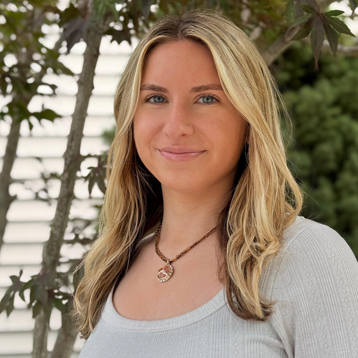 May include: A light gray ribbed long-sleeve shirt paired with a beaded necklace featuring a round pendant with a textured, metallic finish. The necklace consists of small, dark brown beads, while the pendant displays a circular, ornate design.